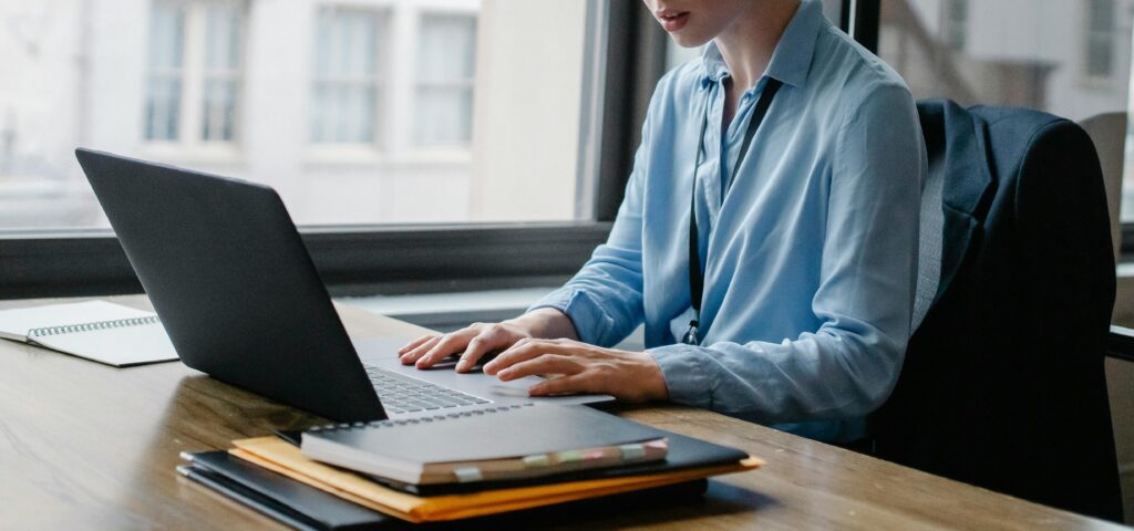 Crop anonymous female secretary surfing internet on netbook at table with agenda and folders near window at work